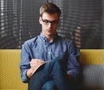 A young man in glasses writes in a notebook while sitting on a stylish couch indoors.