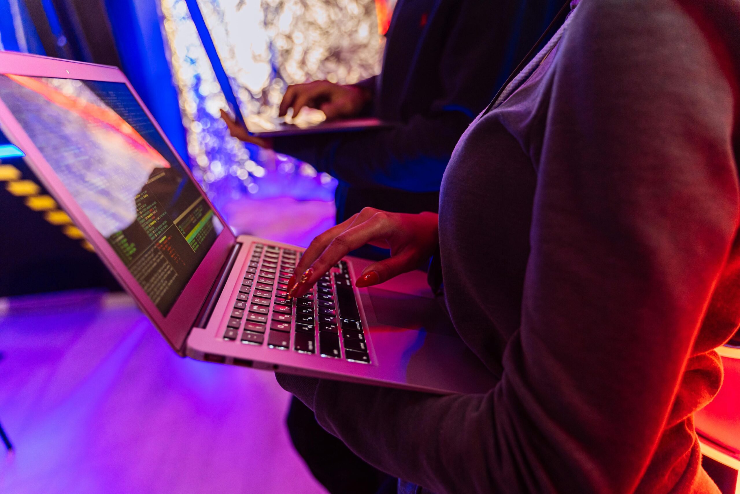 Two professionals analyzing data on laptops with colorful lighting, focusing on cybersecurity details.